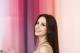 A woman with long brown hair standing in front of a pink wall.